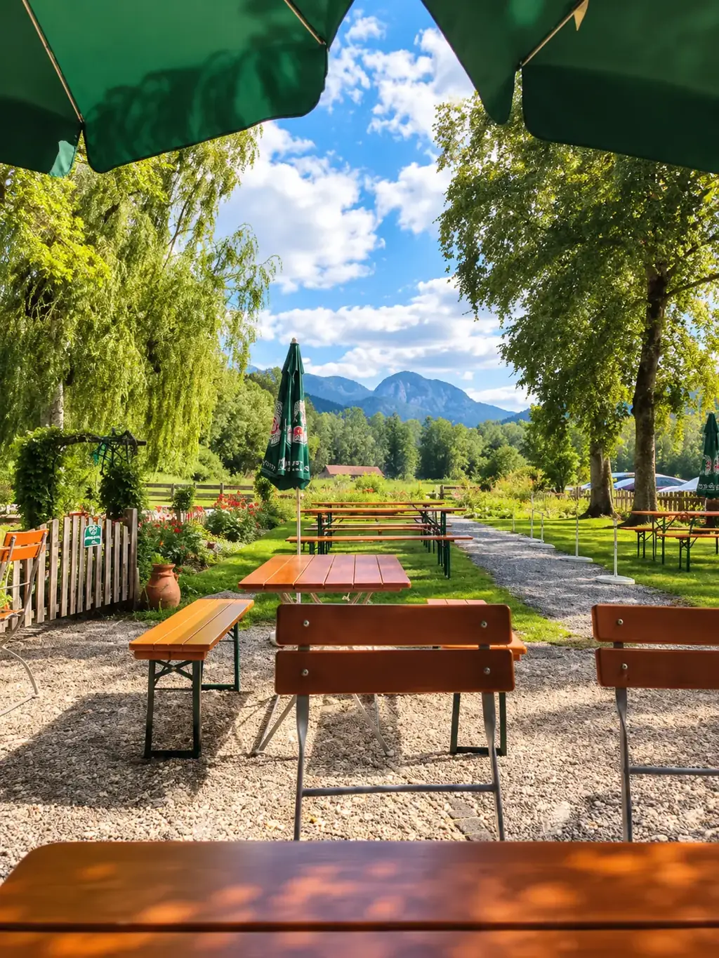 Blick aus dem Biergarten unter grünen Schirmen über Tische und Kiesweg auf die Berge