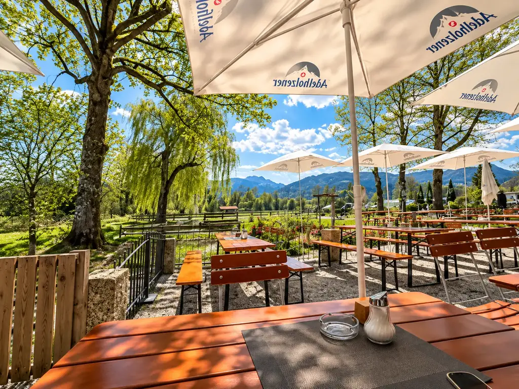 Biergarten mit weißen Sonnenschirmen unter alten Bäumen, dahinter eine Trauerweide und die Berge
