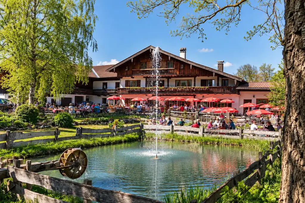 Die Moosmühle mit Biergarten und roten Sonnenschirmen, davor der Teich mit Fontäne und einem kleinen Wasserrad